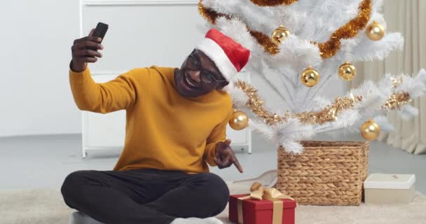 Foto de Año Nuevo, hombre afroamericano estudiante negro se sienta cerca del árbol de Navidad en casa en el suelo en la sala de estar utiliza el teléfono móvil para crear foto selfie toma video de vacaciones para amigos en línea en la cámara — Vídeos de Stock