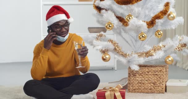 Afro amerykański facet czarny człowiek siedzi pod choinką nosi czapkę Mikołaja i medyczna maska ochronna pije szampana ze szkła mówi przez telefon wita zdalnie szczęśliwy Nowy Rok, epidemia koronawirusów — Wideo stockowe