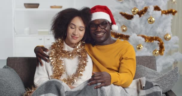 Young couple afro american family boyfriend and girlfriend curly woman and black man wears winter sweaters sitting together on sofa home against background of Christmas tree hugging looking at camera — Stock Video