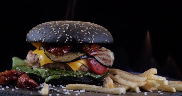 O hambúrguer preto com carne e verduras com batatas fritas está na mesa contra o fundo do fogo. Chisburger com salada de tomate molho de cebola e carne bovina e pão escuro na grelha cercada por chamas prato quente — Vídeo de Stock