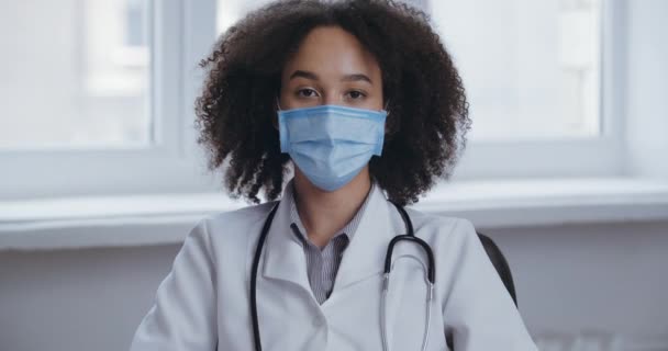 Femme aux cheveux bouclés médecin infirmière confiante ou étudiante en médecine porte un masque chirurgical sur son visage, s'assoit à table, utilise une webcam pour communiquer avec les patients, consulte en ligne à distance via un réseau — Video