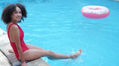 Aktif, kıvırcık Afrikalı esmer, havuz başında şişme çöreklerle oturuyor, mavi su havuzunda sallanan bacaklar, serinliyor, serinliyor, oynuyor, sallanıyor, sıçramalar ve dalgalar yaratıyor, neşeli bir şekilde kameraya bakıyor.