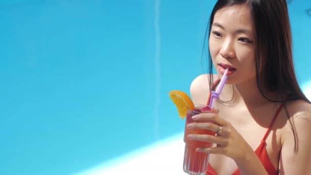 Retrato de uma jovem mulher asiática desfrutando de dia quente de verão, tomando sol bebendo refrescante coquetel alcoólico através de palha, relaxa perto da piscina em resort exótico no exterior, prazer, câmera lenta — Vídeo de Stock