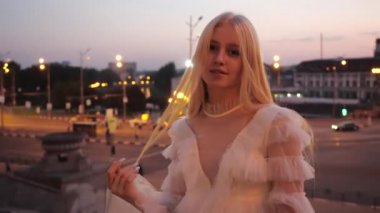 Beyaz elbiseli sevimli sarışın kız uzun ipeksi saçlarını yudumluyor ve akşam şehir merkezinin arka planına gülümsüyor, arabalar ve ışıklar, Harkov, Ukrayna. Güzel sarışın parkta yürümekten hoşlanıyor..
