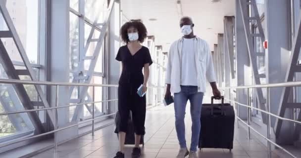 Etnische Afro-Amerikaanse echtpaar dragen beschermende medische maskers en het dragen van koffers wandelingen in de luchthaven terminal. Vrouw met afro kapsel en zwarte man die bagage vervoert tijdens het coronavirus — Stockvideo