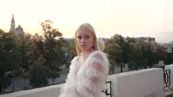 Cinematic side view of a wonderful young european blonde in a stylish white dress effectively walking outside along an observation deck amid trees, a road and a church. Girl walk around the city. — Stock Video