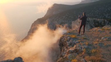 Dağın eteğindeki renkli bulutların üzerinde duran turist kız akıllı telefonuyla fotoğraf çekiyor. Uçurumun kenarında bir kadın gün batımını ve buharlaşmayı filme alıyor.