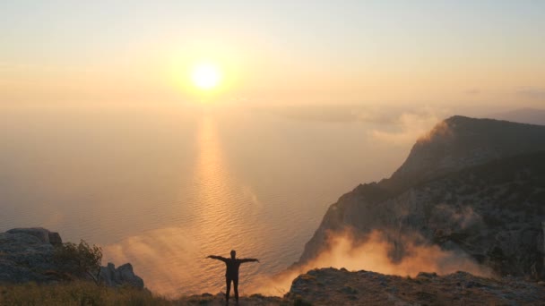 Silhouette of a girl standing on the edge of a high mountain and enjoying a beautiful bright sunset over the sea. Active athletic woman spreads her arms to the sides and imagines that she is a bird. — Stock Video