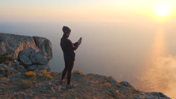 Disparo circular desde la espalda de una chica parada en el borde de un acantilado que toma vídeo y fotos con smartphone de hermoso humo y un par de nubes sobre el telón de fondo de la puesta del sol y el mar. — Vídeo de stock