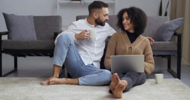 Genç bir aile, oturma odasında oturmuş dizüstü bilgisayara bakıyor, e-postaları okuyor, internetten video izliyor, beyaz bardaklardan kako kahvesi içiyor.