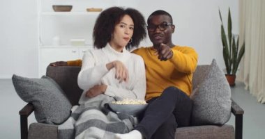 Mutlu Afro-Amerikan çifti genç aile erkek arkadaşı ve kız arkadaşı kanepede oturup TV programları izleyip kamera tebessümlerine parmak işareti yaparak patlamış mısır yiyor.