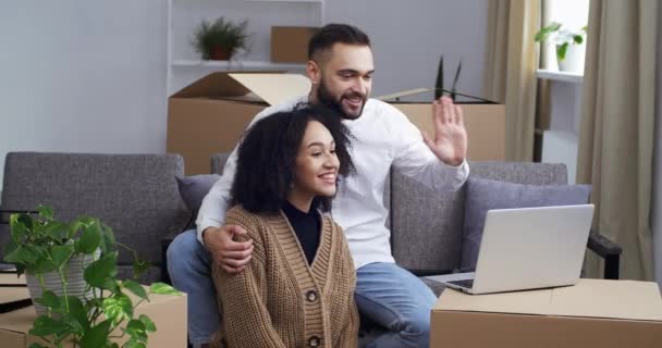 Feliz pareja multiétnica caucásico hombre y mujer africana haciendo videoconferencia en línea con ordenador portátil después de la reubicación. Amigos saludo familiar mostrando nueva casa y cajas charlando en red — Vídeos de Stock