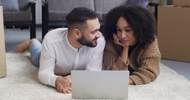 Close-up multiethnic para kręcone czarny kobieta i biały mężczyzna leżą na dywanie na podłodze domu pokój dzienny za pomocą laptopa do komunikowania się z kamerą internetową, rozmawiając z przyjaciółmi fale pozdrowienia strony — Wideo stockowe