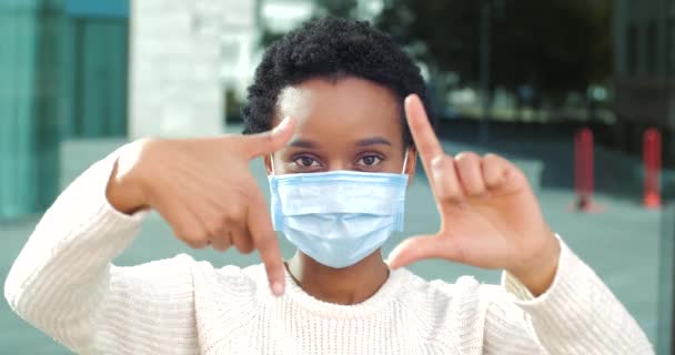 Афро-американская девушка в медицинской защитной маске стоит на улице, глядя в камеру, кладет руки перед собой, делает прямоугольную форму пальцами, указывая на глаз, делая вид, что фотографирует. — стоковое видео
