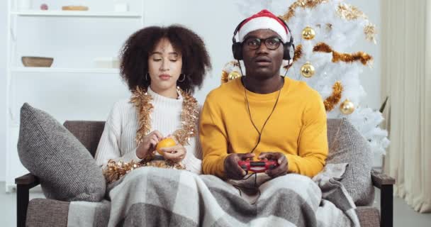 Karışık ırk çifti Afro-Amerikan erkek ve kadın evdeki oturma odasında kanepede birlikte oturuyorlar. Mandalina yiyen kıvırcık genç kız dargınlıktan gücenmiş. Konsol oynayan arkadaşına bakıyor. — Stok video