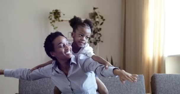 Αφρο-αμερικανική μαύρη γυναίκα μαμά με την κόρη προσποιούνται ότι είναι αεροπλάνο, ενώ κάθεται στον καναπέ στο σαλόνι. Μητέρα και χαριτωμένο θηλυκό παιδί παιδί παίζουν το παιχνίδι διασκεδάζοντας στο σπίτι περνούν το χρόνο τους μαζί γέλιο — Αρχείο Βίντεο