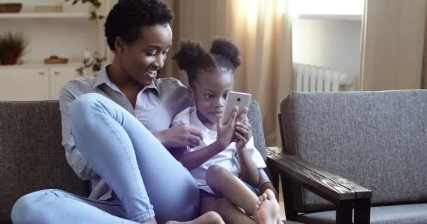 Jovem mãe com curto afro penteado abraços pequena filha curioso misto raça criança negra menina mostra ensina como usar dispositivos móveis smartphone, família tira vídeo fazendo foto sentado no sofá — Vídeo de Stock