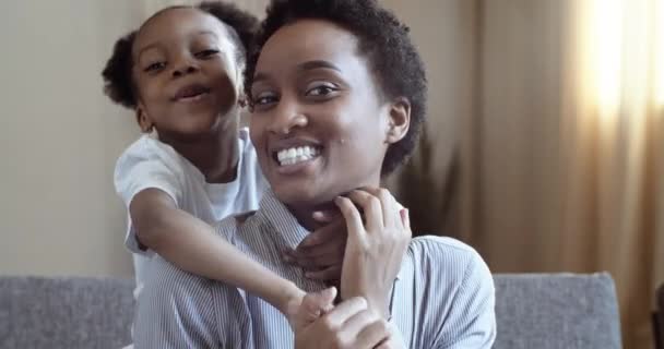 Portret szczęśliwej afroamerykańskiej rodziny. Zdjęcie głowy kochający czarny kobieta młoda matka siedzi na kanapie mało słodkie dziecko dziewczyna przytula rodzica po szyi od tyłu koncepcja przytula do mamy — Wideo stockowe