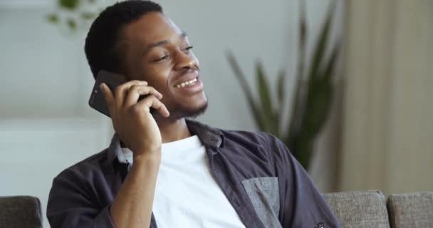 Ritratto di uomo nero sorridente felice volto maschile africano ragazzo americano seduto a riposo si rilassa a casa sul divano parla su smartphone comunica, freelancer lavora a distanza sul telefono facendo affari online — Video Stock
