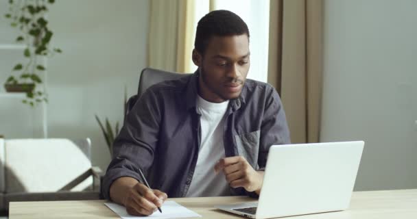 Портрет умный сфокусированный студент черный бизнесмен афро американец парень сидит за столом в домашнем офисе глядя на экран ноутбука делает заметки пишет на бумаге делает тест обучения дистанционно изучения языка — стоковое видео
