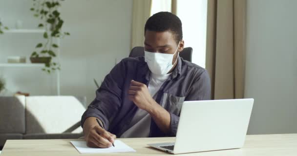 Junge attraktive Afroamerikaner in medizinischer Schutzmaske schreibt Notizpläne macht Berechnungen zeichnet Grafik in Notizbuch auf Papier Blick in Laptop-Bildschirm Studien arbeitet online in Netzwerk aus der Ferne — Stockvideo