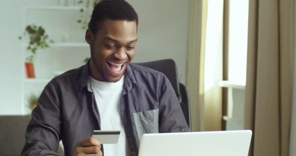Heureux afro-américain gars noir homme d'affaires étudiant assis à table dans le bureau à domicile tenant carte bancaire de crédit dans sa main fait achat en ligne sur Internet à l'aide d'un ordinateur portable se réjouit de réductions vente — Video