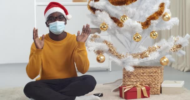Веб-камера зрения молодой черный парень афроамериканец в очках в желтом свитере и Санта шляпа сидит возле елки дома носит медицинскую защитную маску говорит активно жесты С Новым годом 2021 — стоковое видео