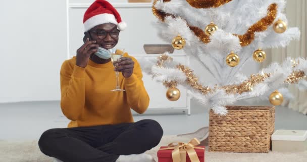 Afro amerykański facet czarny mężczyzna nosi maskę medyczną siedzi w pobliżu choinki w Santa kapelusz trzymając szklankę szampana w rękach mówi przez telefon życzenia smartfona szczęśliwy nowy rok do przyjaciół rodziców zdalnie — Wideo stockowe