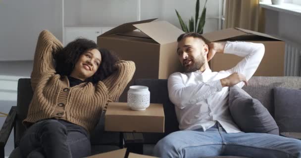 Close-up multi-étnico casal encaracolado afro-americano menina e caucasiano cara sentado no sofá com as mãos dobradas atrás de cabeças descansando relaxante olhando para caixas de papelão com coisas, conceito em movimento — Vídeo de Stock