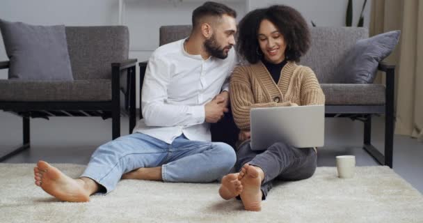 Interracial relationship, african american woman and caucasian man husband and wife sitting at weekend at home on living room floor together looking at laptop having rest checking mail online chatting — Stock Video