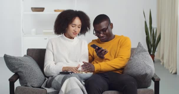 Giovane afro americano coppia uomo e donna seduti insieme sul divano in soggiorno a casa mangiare popcorn ridere divertirsi, ragazzo nero guardando smartphone cellulare chat online tiene gadget — Video Stock