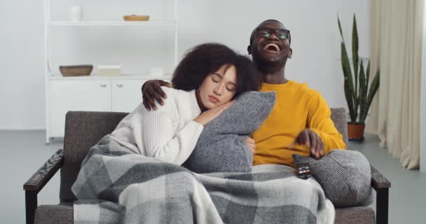 Black man laughs loudly smiles toothy watching comedy film program tv switches channel hugs sleeping tired girl wife afro american woman with curly hair covered with blanket sitting together at home — Stock Video