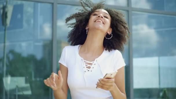 Amerikaanse Afrikaanse etnische vrouw dansend haar handen opsteken met muziek die smartphone in de straat houdt. Gelukkig krullend meisje met vrolijke emoties van vreugde houdt van het leven en outdoor activiteiten te genieten, slow motion — Stockvideo
