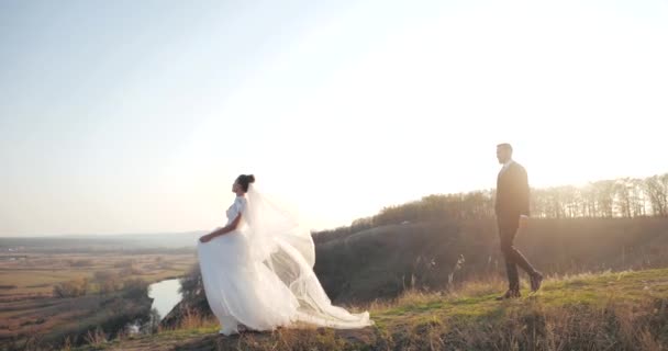 Belle mariée en robe blanche luxueuse vient au bord de la colline et admire le vaste paysage, suivie par son marié barbu en costume. Couple de mariage européen marchant dans une zone déserte à l'automne. — Video