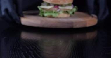 Yakın planda kesilmiş siyah lastik eldivenli, yemek pişirmek için özel üniformalı bir erkek eli kafe restoranında hamburger, et ve sebzeli hamburgerle ahşap kaplama servis ediyor.