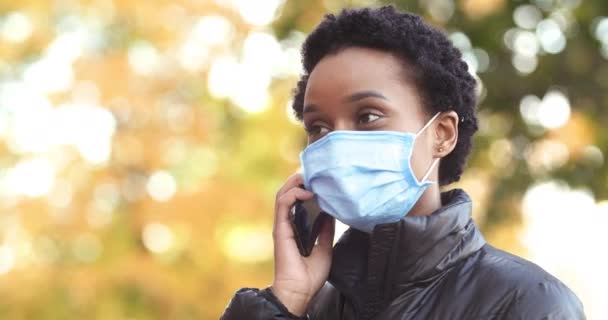 머리에는 의료 마스크를 쓴 아프리카계 미국인젊은 여성이 핸드폰으로 이야기하며 야외에 서 있는 모습이 찍혔다. 호흡기 보호를 받는 행복 한 여성 사업가가 핸드폰으로 말하는 모습 — 비디오