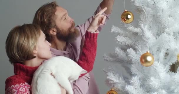家族の女の子と男白い猫ペットと2人の友人一緒にお祝いの装飾で家を飾る休日の準備のための新年を祝う、男はクリスマスツリーのための黄金のボールを保持し、ホームパーティー — ストック動画