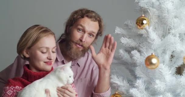 Close-up retrato branco casal mulher da família com cabelo curto e barbudo marido e mulher segurando gato branco brincando com pet mostra decorações do ano novo acenando Olá mostrar polegar como na câmera — Vídeo de Stock