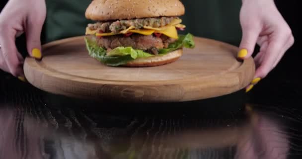 Close-up kobieta szef kuchni kelner serwuje hamburgery na drewnianej desce stawia fast food na stole. Przycięta rama żeńskich rąk z żółtym manicure prezentują pyszne apetyczny cheeseburger z sałatką pomidorową — Wideo stockowe