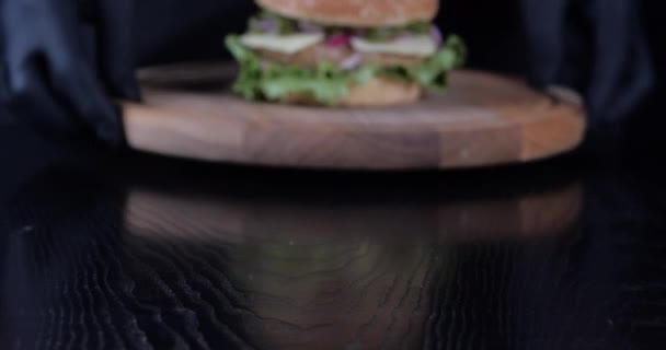 Крупным планом обрезанный снимок мужских рук в черных резиновых перчатках в специальной униформе для приготовления пищи подает гамбургер на столе в кафе-ресторане аппетитный бургер с мясом и овощами крутит деревянную доску — стоковое видео