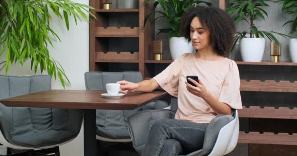Wanita bisnis hitam gadis keriting cantik muda minum kopi dari cangkir putih menikmati teh cappuccino kakao duduk di kafe dengan alat modern telepon menanggapi obrolan pesan santai di restoran — Stok Video