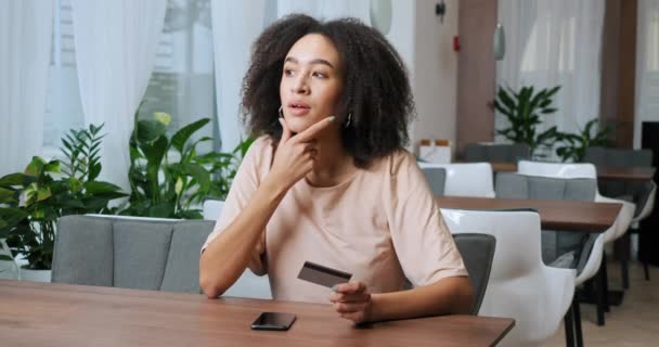 Primer plano del visitante afroamericano de café chica rizada mujer con tarjeta de crédito de plástico en sus manos y teléfono inteligente gadget móvil moderno hace la compra en línea en Internet compra cuidadosamente regalo — Vídeo de stock