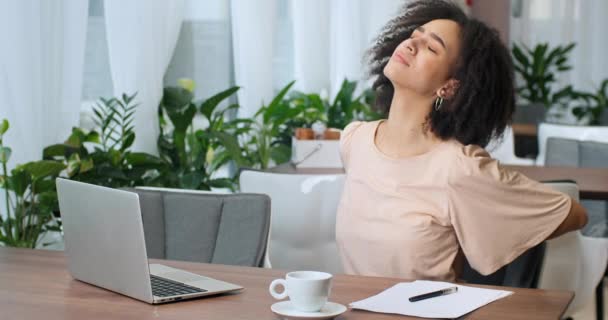 La mujer afroamericana de pelo rizado freelancer sentada en un restaurante cafetería trabajando remotamente desde un lugar público siente dolor sufre de lesión en la artritis de espalda aferrándose a la columna vertebral, problemas de enfermedad — Vídeos de Stock