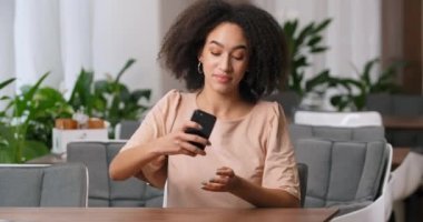 Güzel bukleli Afro-Amerikan kadın portresi bej bluz giyer evdeki koltukta oturur ya da telefon oyununda oyun oynar akıllı telefon kazanır sevinçle ellerini kaldırır.