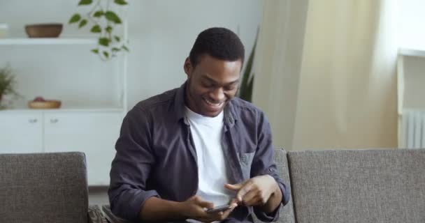 Misto raça cara afro americano preto homem sentado em casa na sala de estar no sofá aprende boa notícia feliz para ganhar loteria sente triunfo assistindo tv chamadas no telefone compartilhar emoções fala no smartphone gadget — Vídeo de Stock