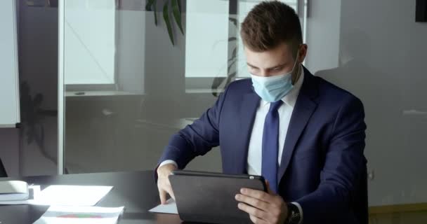 Retrato hombre de negocios en traje sentado en la mesa en la oficina lleva máscara médica protectora contra la infección virus respiratorio se ve en la computadora tableta dispositivo lee contrato documento analiza finanzas — Vídeo de stock