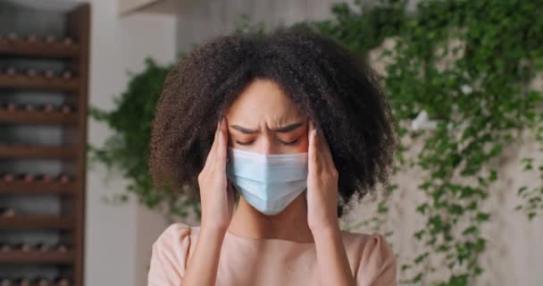 Vista frontal de mujer afroamericana joven niña lleva mascarilla durante pandemia que sufre de dolor de cabeza siente síntomas de migraña espasmo sostiene la frente con las manos frotando sus sienes coronavirus — Vídeo de stock