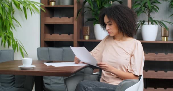 Mujer concentrada estudiante afroamericana freelancer con el trabajo de papel sentado en la mesa en la oficina restaurante tiene hojas blancas de papel en sus manos documento aviso lee cartas dadas informes — Vídeo de stock