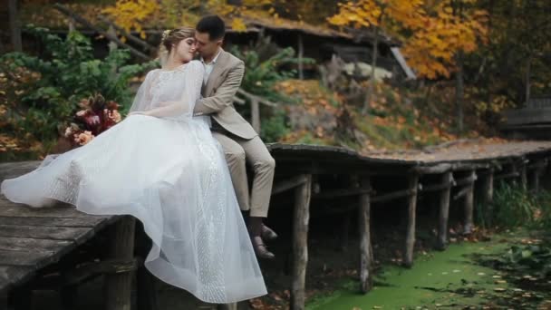 Schöne junge Hochzeitspaar umarmt und streichelt, während sie auf einer Holzbrücke am See vor dem Hintergrund der Herbstblätter sitzen. Braut im Kleid und Blumenstrauß mit dem Bräutigam. — Stockvideo