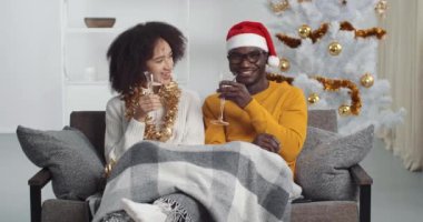 Happy afro american couple clink champagne glasses toast and celebrate together New 2021 Year at home in living room sitting against glowing Christmas tree, holiday party. Congratulations concept